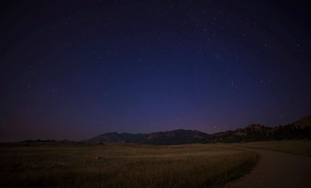 A starry sky over a grassy field and mountains.