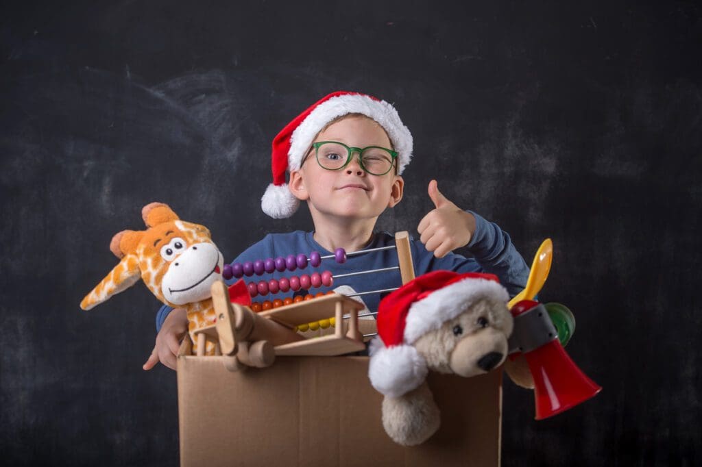 A boy in a santa hat and santa claus hat is holding toys in a cardboard box.