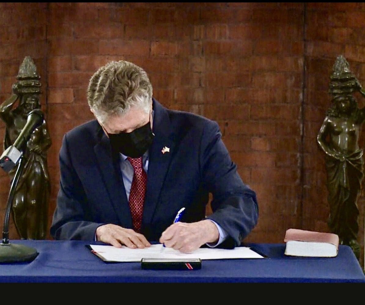 A man wearing a mask signing papers at a desk.
