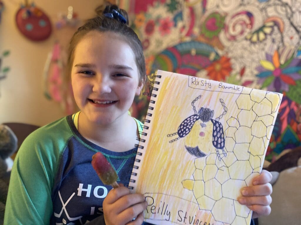 A girl holding up a notebook with a drawing of a bee.