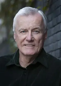 An older man smiling in front of a brick wall.