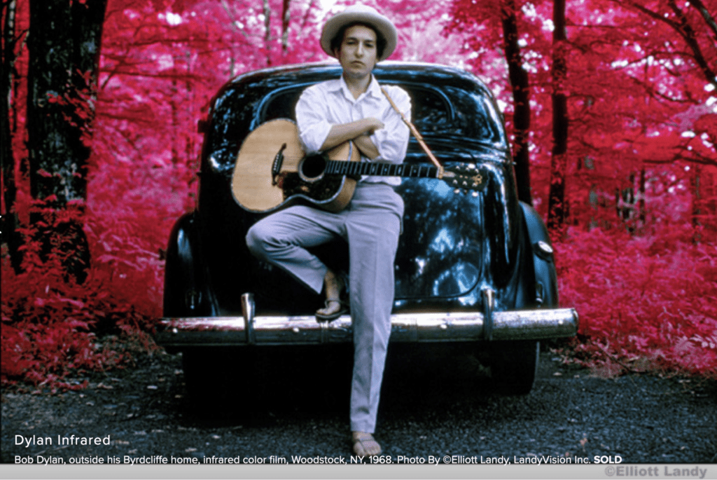 A man with a guitar leaning on the back of a car.