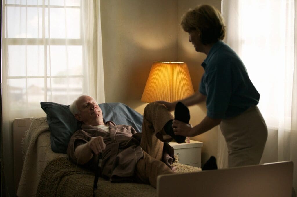 A woman helping an elderly man to his bed.