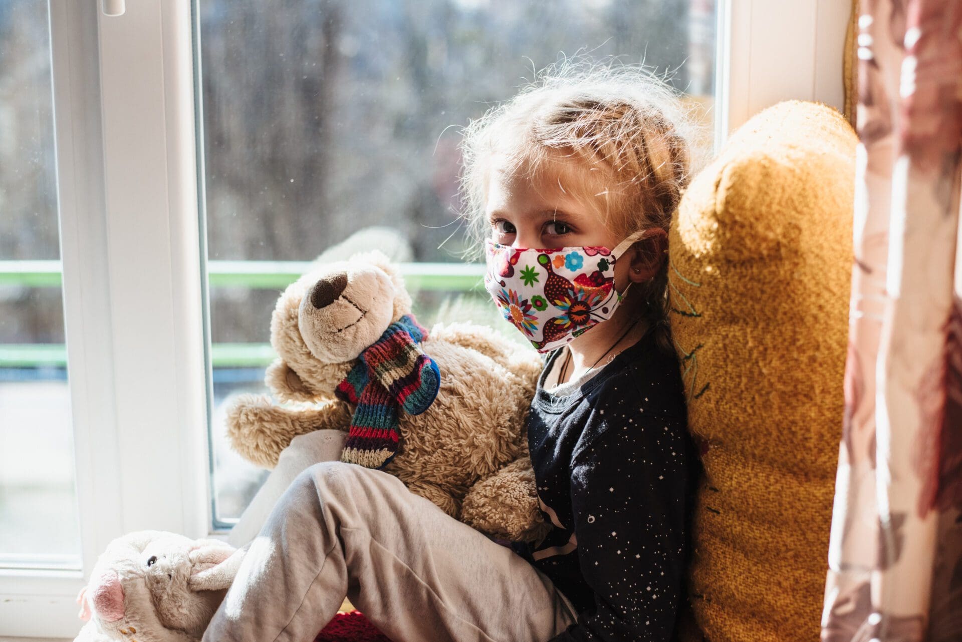 A little girl wearing a face mask sitting on a window sill with a teddy bear.
