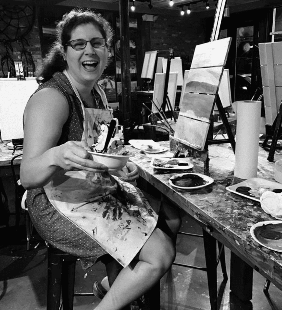 A woman in a black and white photo smiling at a painting table.
