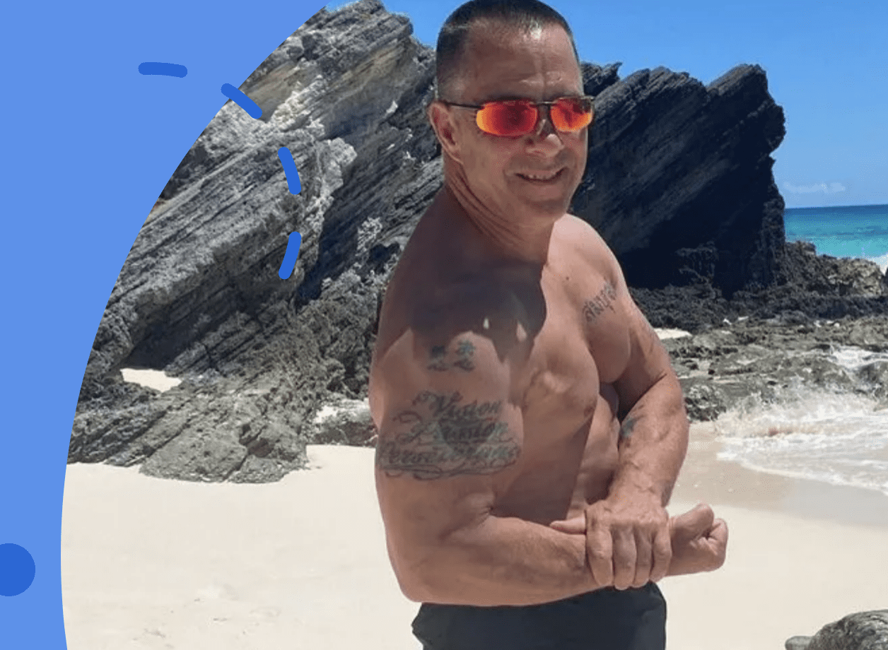 A shirtless man posing in front of a rocky beach.