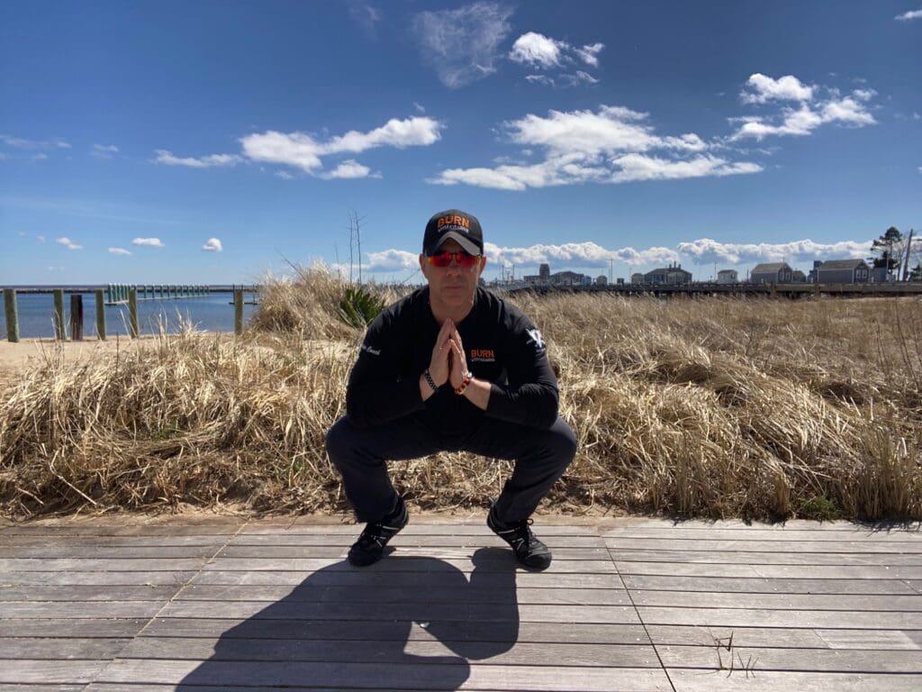 A man squatting on a boardwalk near the ocean.