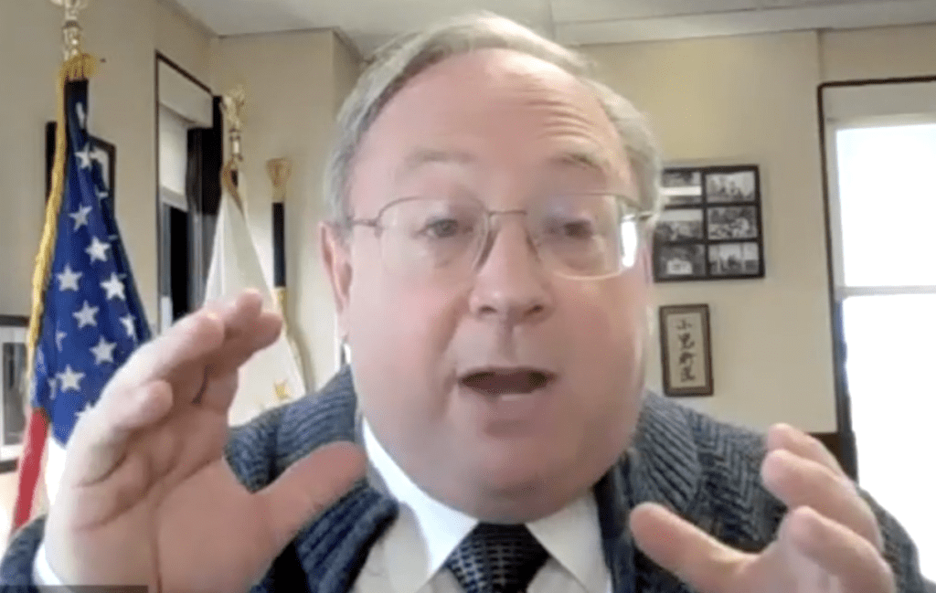 A man wearing glasses and a tie is speaking into a camera.