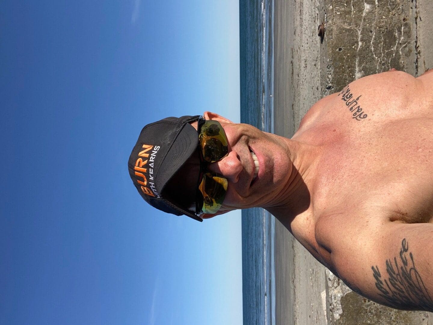 A man in a shirt and hat taking a selfie on the beach.