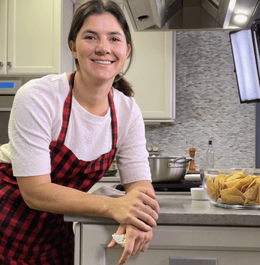 A woman in an apron smiling in a kitchen.
