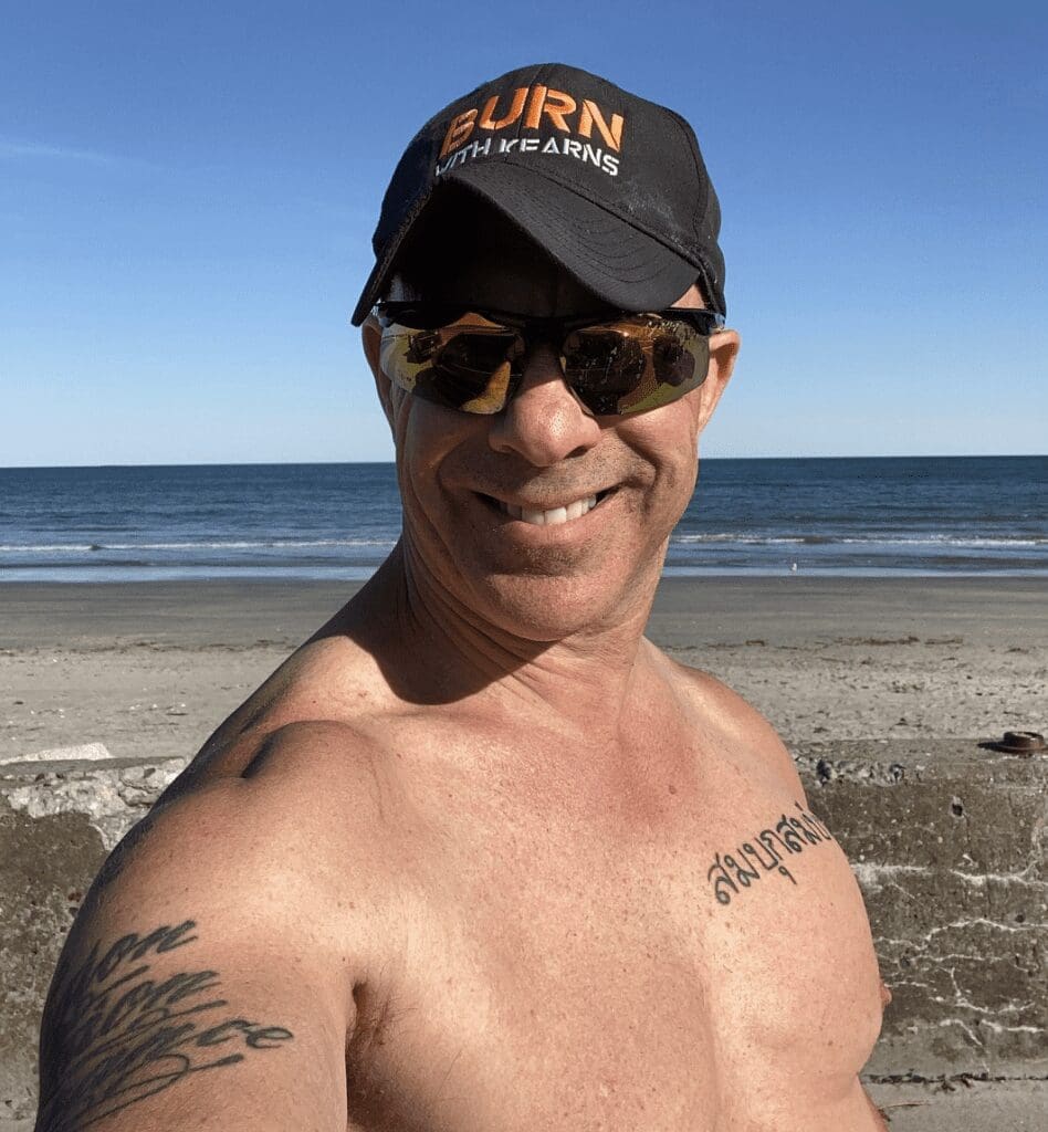 A man with tattoos and a hat in front of the ocean.
