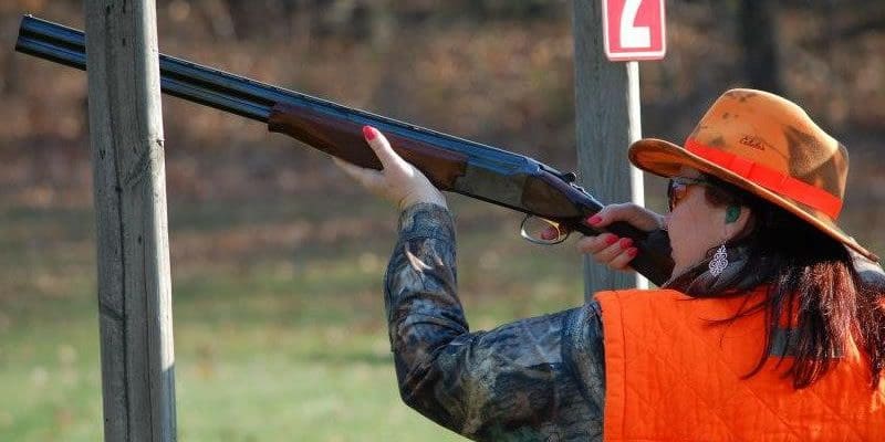 A woman wearing an orange hat and orange vest shooting a shotgun.