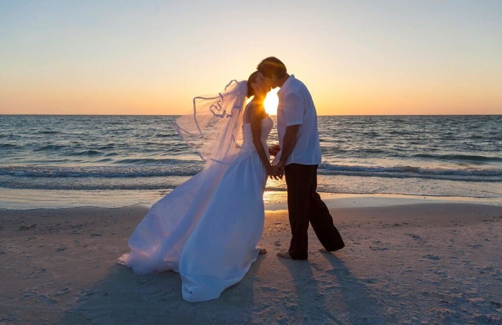 A bride and groom kissing on the beach at sunset.