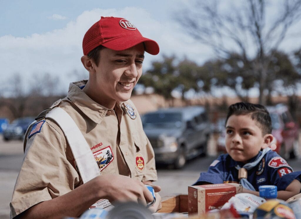 A boy in a scout uniform is standing next to a box of food.