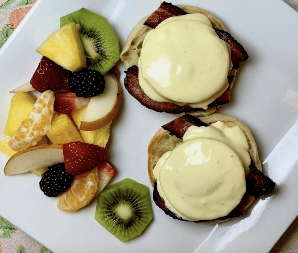Two eggs benedict with fruit on a white plate.