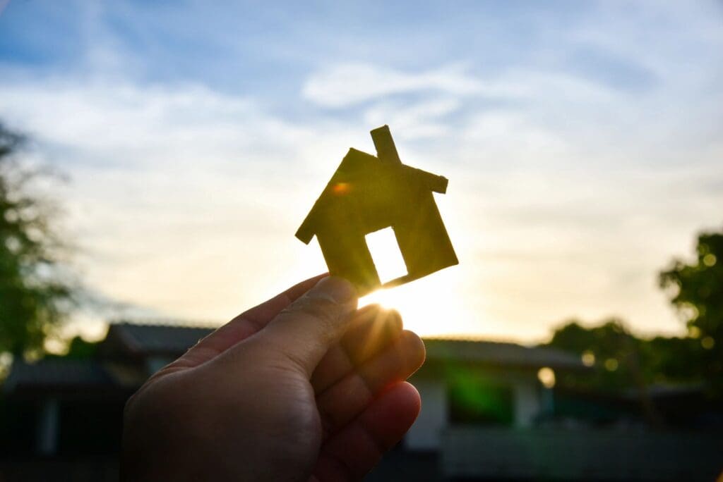 A person holding a house model in front of a sunset.