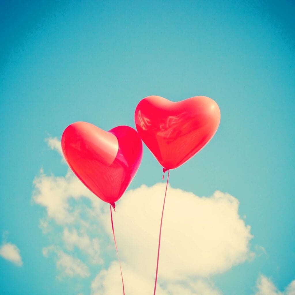 Two red heart shaped balloons against a blue sky.