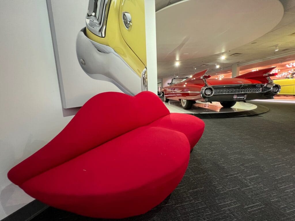 A red couch sits in front of a car in a museum.