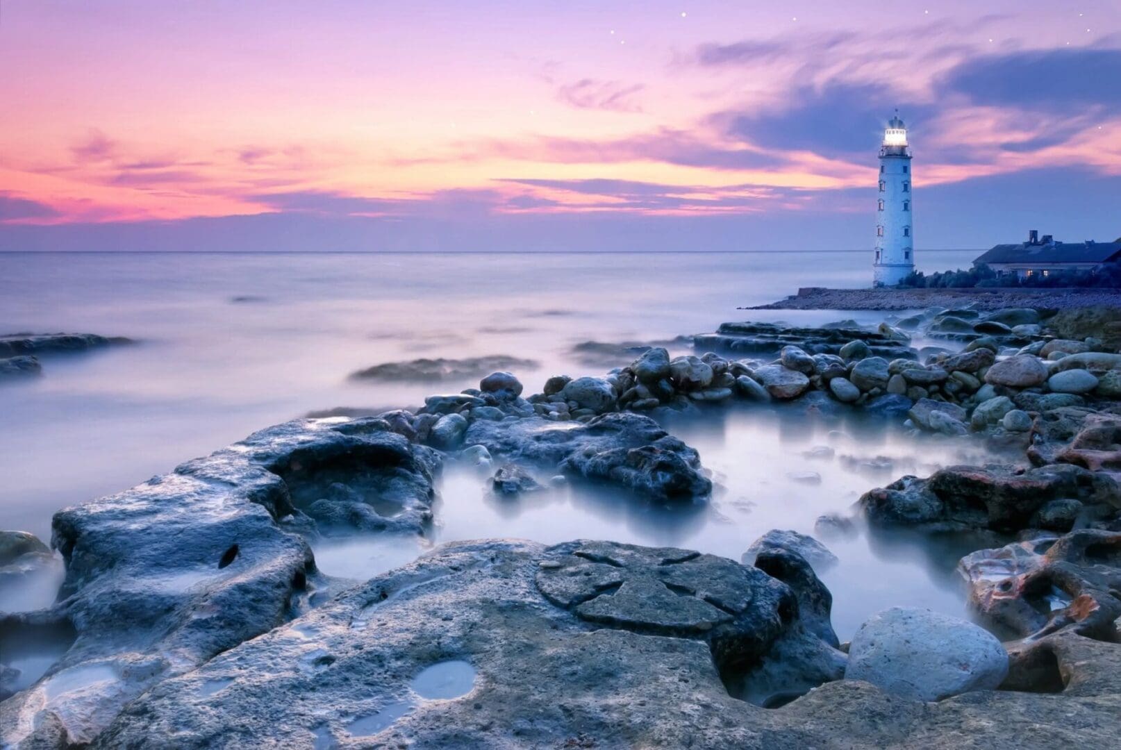 A lighthouse sits on a rocky shore at sunset.