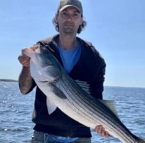 A man holding up a striped bass during an outdoor sports adventure in Rhode Island, on a boat.