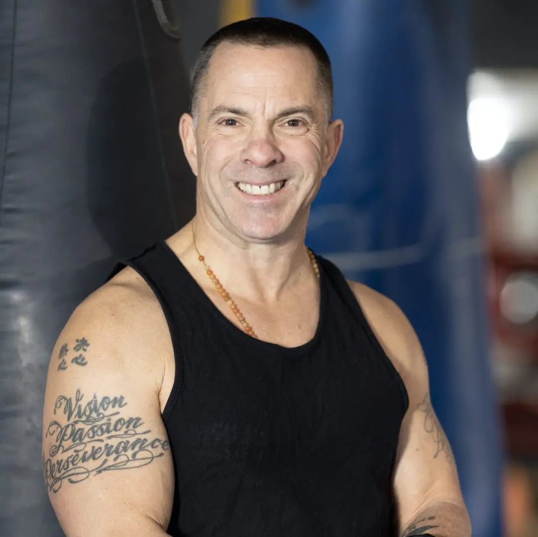 A man in a black tank top with tattoos posing in front of a punching bag.