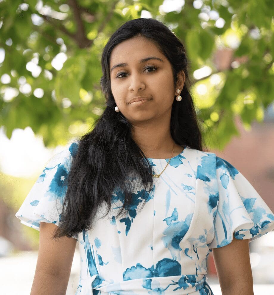 A young woman who identifies as BIPOC in a blue and white floral dress standing in front of a tree.