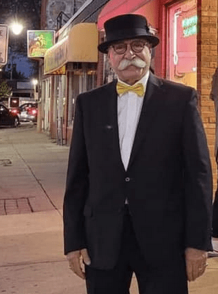 A homeless man in a tuxedo and bow tie standing on a city street.