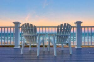 Two adirondack chairs on a deck overlooking the ocean.