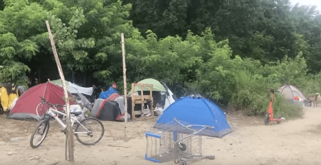 A group of tents and bicycles are parked in a dirt area occupied by homeless individuals.