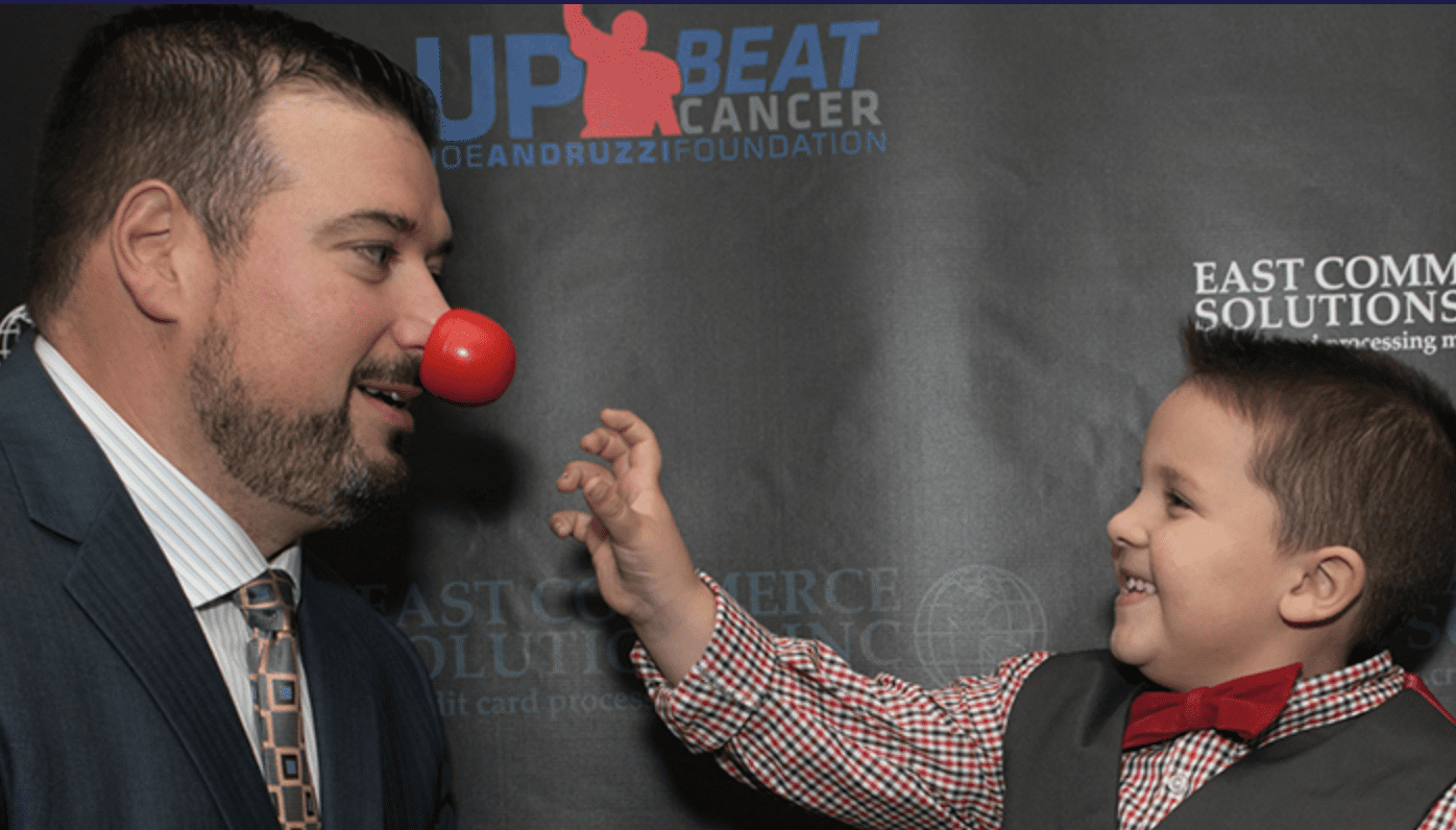 A man in a suit and tie alongside a little boy wearing a red nose, representing the Joe Andruzzi Foundation's philanthropic efforts.