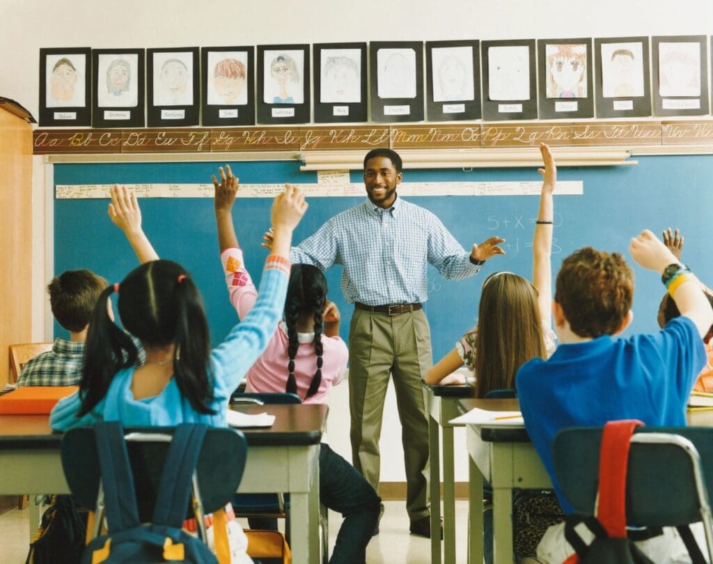 A teacher in a classroom with students raising their hands.
