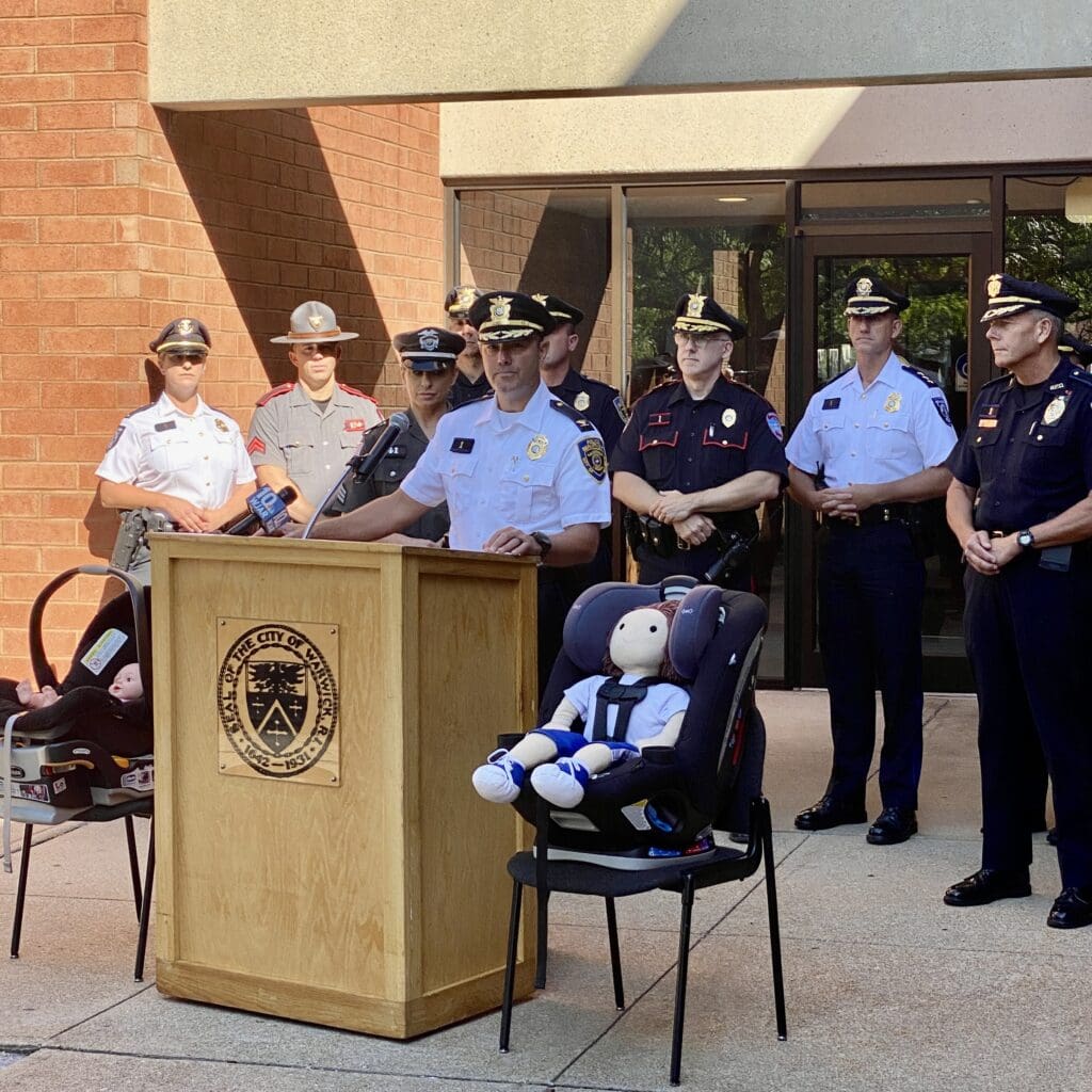 A group of police officers standing in front of a podium discussing car seat safety.
