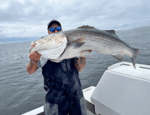 A man proudly displaying a massive striped bass while out on a boat in the picturesque outdoors of Rhode Island.