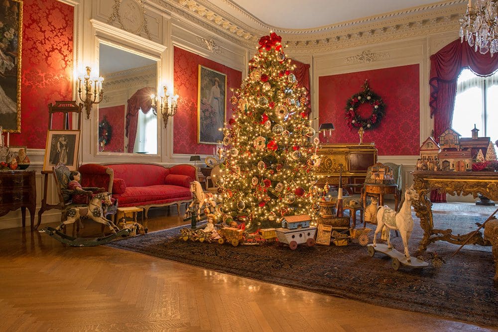 A Christmas tree adorned with sparkling lights and ornaments stands tall in a room with vibrant red walls. The festive atmosphere exudes the charm of Newport mansions during the Christmas season.
