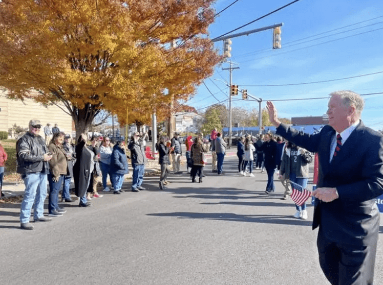 A man in a suit is waving to a crowd of people at the Cranston Veterans Day Parade.