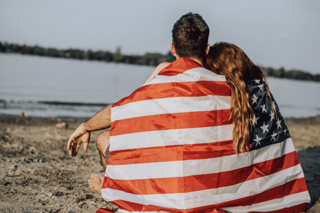 A patriotic couple embracing on the beach, enveloped in an American flag.