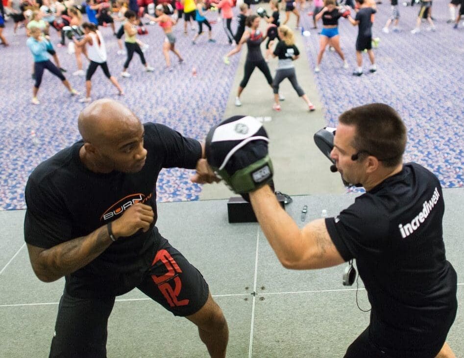 Two men engaged in a self-defense workout session at a gym.