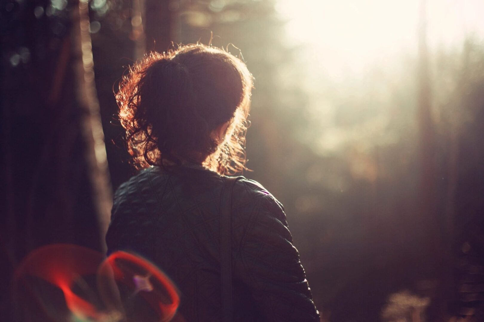 A woman is standing in the woods looking at the sun.