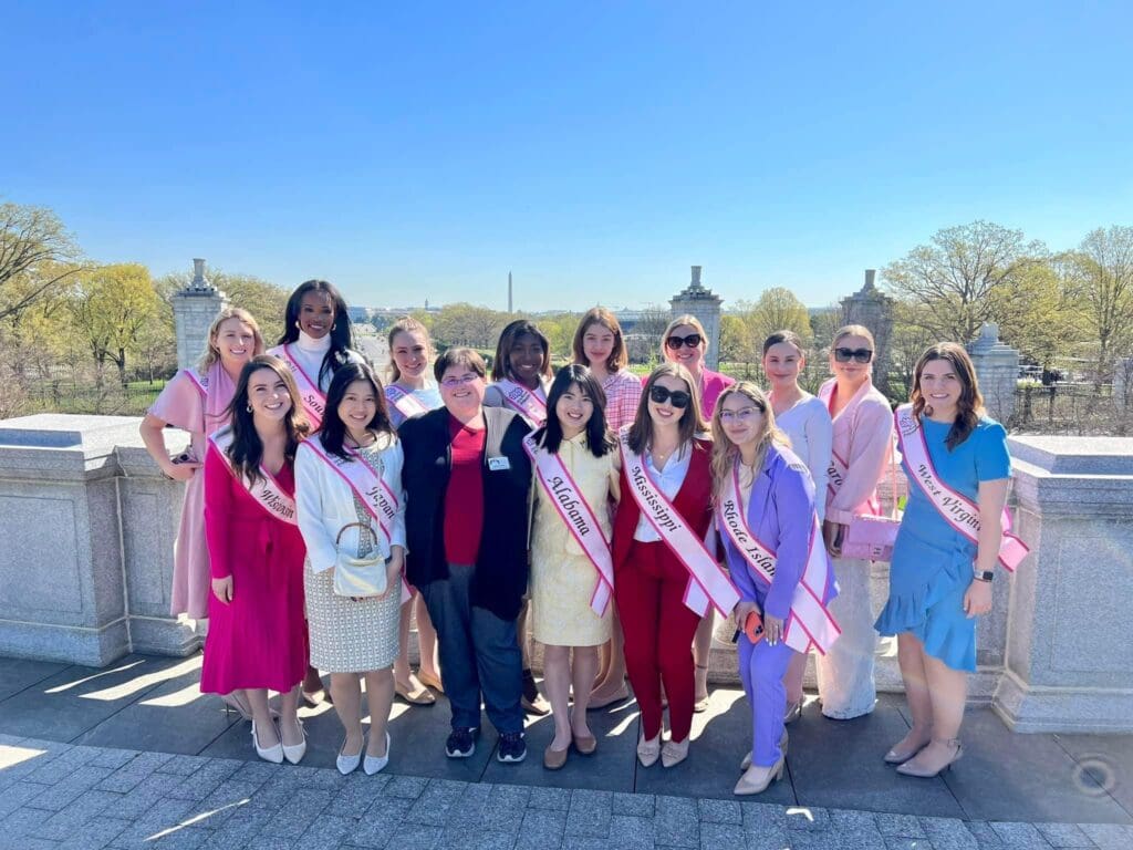 A group of women in pink sash posing for a photo under cherry blossoms.