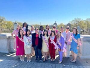 A group of women in pink sash posing for a photo under cherry blossoms.