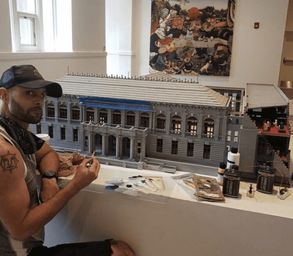 A man sitting in front of a legos model of a building.