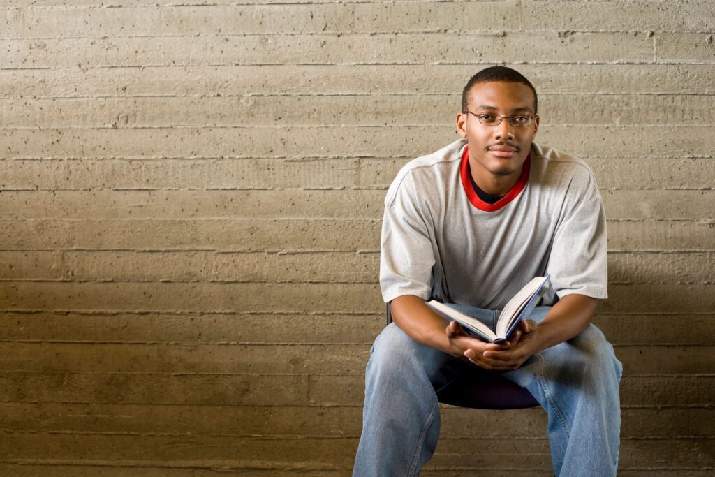 A young black man sitting on a chair reading a book.