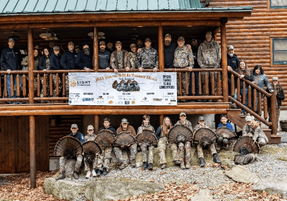 A group of people posing in front of a cabin before a turkey shoot for youth.