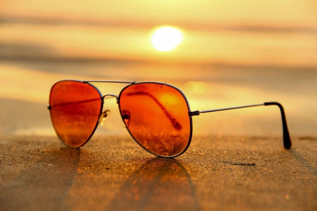Orange-tinted sunglasses resting on a sandy beach at sunset.