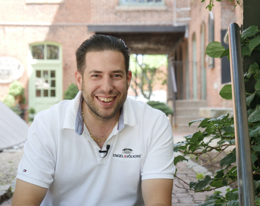 Smiling man in a white polo shirt outdoors near greenery and brick buildings.