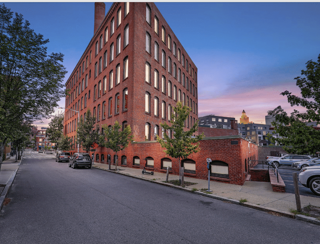 Brick building on a street corner during sunset with trees and parked cars.