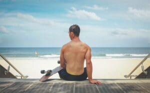 Man sitting on a boardwalk facing the ocean with a skateboard beside him.