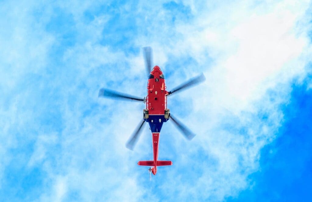 Red helicopter flying against a bright blue sky with clouds.