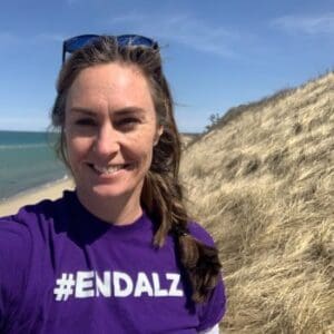 A woman wearing a purple #ENDALZ shirt stands on a sandy dune by the ocean.