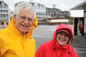 Two elderly people smiling outdoors in bright jackets near water.